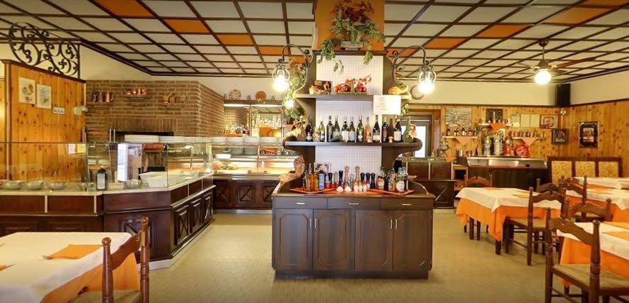 La sala della pizzeria: bancone in legno scuro, bottiglie, tovaglie arancioni e il muro in mattoni del forno